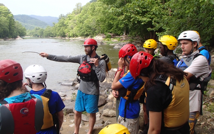 A group of students wearing safety gear watch as an instructor uses a stick to point out over a river.